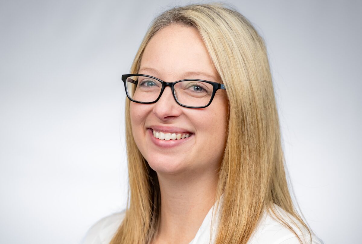 Megan is looking at the camera and can be seen from her shoulders up with white shirt and gray background background